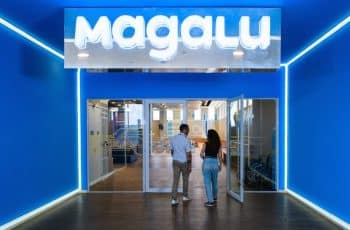 Fachada da Arena Magalu, em São Paulo. Paredes azuis e placa na parte superior com os dizeres "Magalu" em led branco. Portas transparentes com duas pessoas entrando