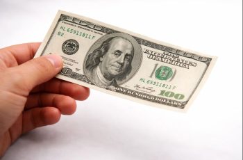 Mão masculina segurando uma nota de 100 dólares, representando a queda do dólar frente ao real no encerramento do ano