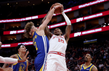 Two basketball players, one in a white uniform and one in a blue and yellow uniform, jump and reach for the ball amid other players on the court.